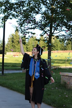A graduate in a black cap and gown celebrates outdoors, surrounded by green trees and grass. The individual is holding a stick from which colorful confetti is being released.