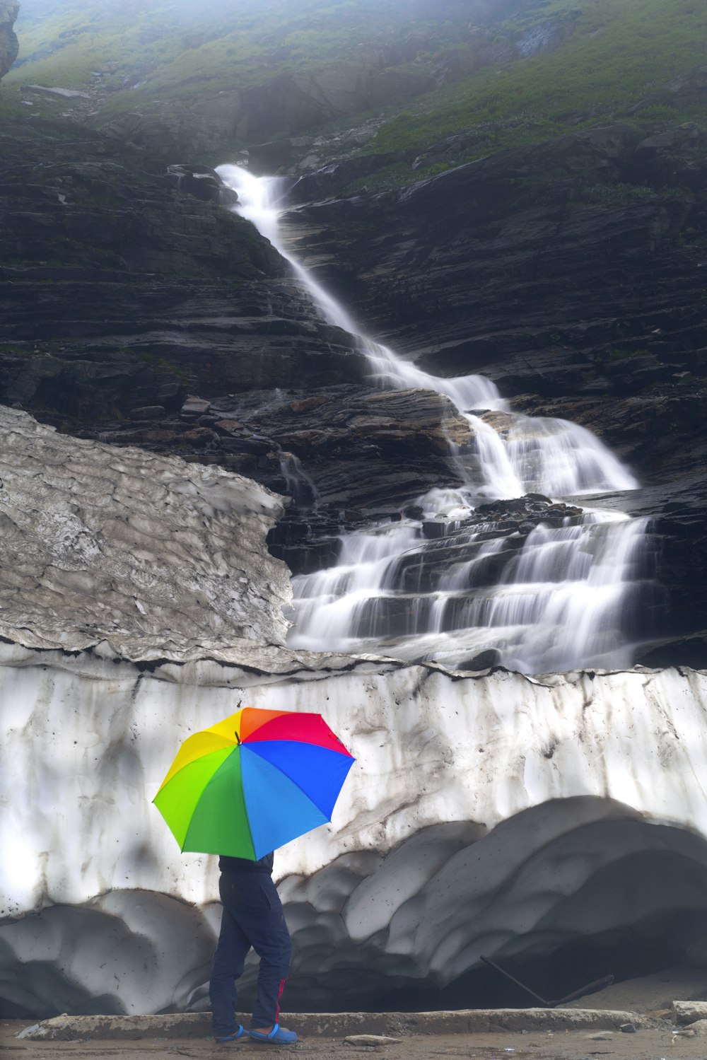person holding umbrella standing on rock formation near waterfalls during daytime