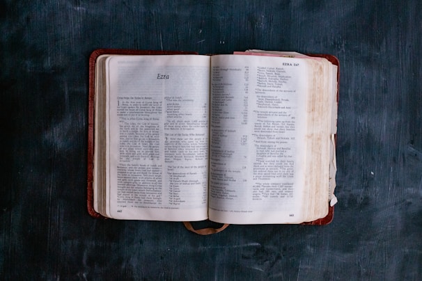 An open book with readable pages lies flat against a textured dark surface. The pages contain text with the heading 'Ezra', indicating a section of the Bible. The book's cover is a deep red and slightly worn at the edges.