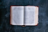An open Bible lies on a dark, textured surface. The pages are open to the Book of 2 Kings, with visible text and chapter headings. The book's cover is a deep red leather, slightly worn, and the edges of the pages are slightly yellowed, suggesting age and frequent use.
