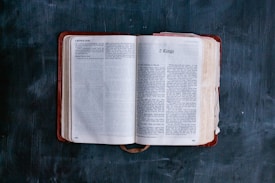 An open Bible lies on a dark, textured surface. The pages are open to the Book of 2 Kings, with visible text and chapter headings. The book's cover is a deep red leather, slightly worn, and the edges of the pages are slightly yellowed, suggesting age and frequent use.