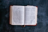 An open book lying on a dark surface, displaying text with the title 'Acts' visible. The pages are slightly aged, with a bookmark ribbon peeking out from the bottom.