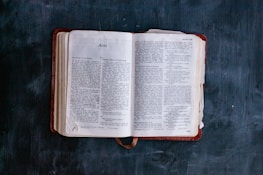 An open book lying on a dark surface, displaying text with the title 'Acts' visible. The pages are slightly aged, with a bookmark ribbon peeking out from the bottom.
