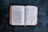 An open book lying on a dark surface, displaying text with the title 'Acts' visible. The pages are slightly aged, with a bookmark ribbon peeking out from the bottom.