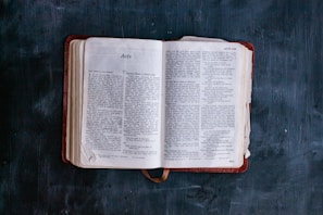 An open book lying on a dark surface, displaying text with the title 'Acts' visible. The pages are slightly aged, with a bookmark ribbon peeking out from the bottom.