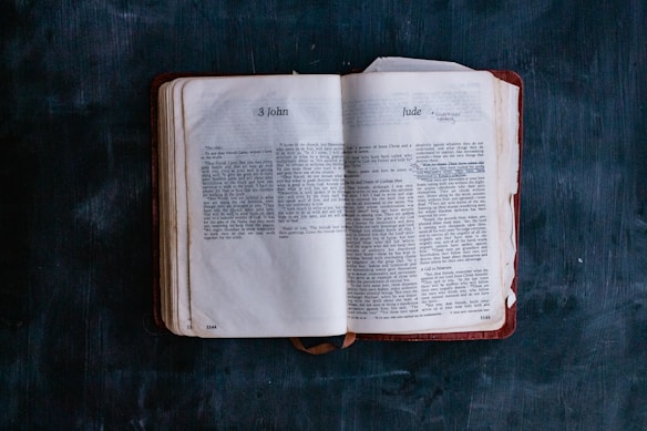 An open book with visible text from the books of 3 John and Jude, positioned on a dark textured background. The pages appear slightly worn, with a red-brown cover visible under the pages.