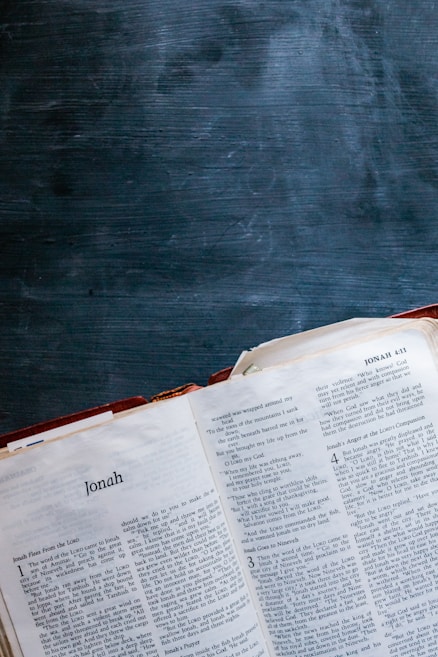 An open Bible displaying the Book of Jonah, with its pages spread and a dark, textured chalkboard surface in the background.