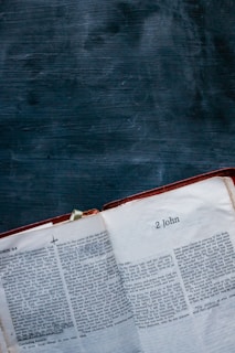 An open book lies on a dark textured surface, displaying pages from the biblical book '2 John'. The text is printed in two columns with chapter and verse annotations visible.