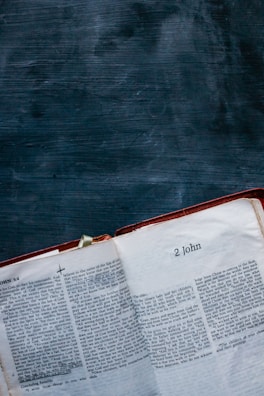 An open book lies on a dark textured surface, displaying pages from the biblical book '2 John'. The text is printed in two columns with chapter and verse annotations visible.