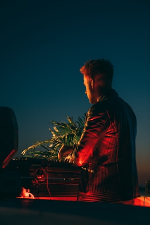 A person wearing a jacket is standing in front of DJ equipment outdoors. The scene is illuminated by warm lighting that contrasts with the darkening sky. Some tropical plants are visible in the background, adding a lively touch.