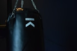 A black punching bag hangs against a dark background. The lighting highlights the text 'OUTSHOCK' on the bag along with an upward pointing arrow. Chains are visible suspending the bag from above, and the scene is dimly lit.