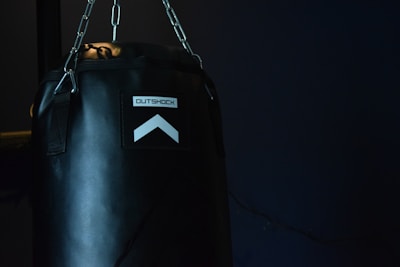 A black punching bag hangs against a dark background. The lighting highlights the text 'OUTSHOCK' on the bag along with an upward pointing arrow. Chains are visible suspending the bag from above, and the scene is dimly lit.