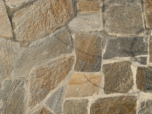 A textured stone wall composed of large, irregularly shaped stones in varying shades of brown, gray, and beige. The stones are fitted together with cement, creating a rugged and natural appearance.