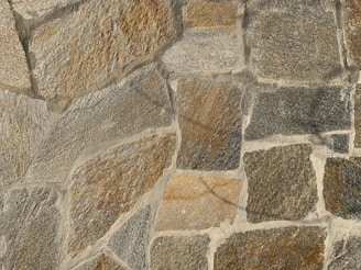 A textured stone wall composed of large, irregularly shaped stones in varying shades of brown, gray, and beige. The stones are fitted together with cement, creating a rugged and natural appearance.