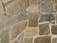 A textured stone wall composed of large, irregularly shaped stones in varying shades of brown, gray, and beige. The stones are fitted together with cement, creating a rugged and natural appearance.