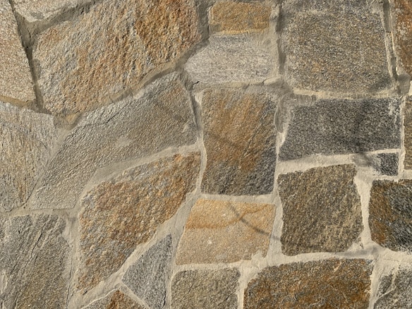 A textured stone wall composed of large, irregularly shaped stones in varying shades of brown, gray, and beige. The stones are fitted together with cement, creating a rugged and natural appearance.