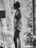 A person wearing a bikini and a robe stands near a large glass window, gazing outside. The background features a blurred view of trees and a balcony, suggesting a serene, natural environment. The black and white tones add a classic and timeless feel to the photograph.