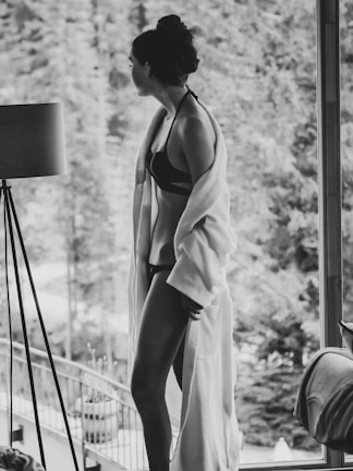 A person wearing a bikini and a robe stands near a large glass window, gazing outside. The background features a blurred view of trees and a balcony, suggesting a serene, natural environment. The black and white tones add a classic and timeless feel to the photograph.