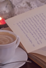 A close-up view of a steaming cup of coffee in a white cup placed on a saucer with a small spoon. Next to it, an open book with handwritten text is visible, and a lit candle in a glass holder provides a warm, cozy ambiance. The background features a lace tablecloth, enhancing the comforting and intimate setting.