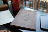 A bar menu with an embossed logo lies on a dark wooden table alongside a phone, a white sheet with text, and a red textured glass. The background features blurred wooden furniture, suggesting an indoor setting.
