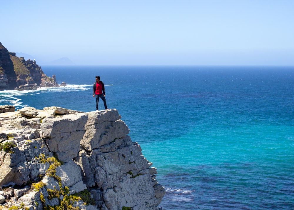 Homme en veste noire debout sur une formation rocheuse grise près de la mer bleue pendant la journée