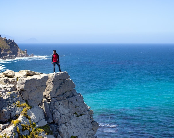 man in black jacket standing on gray rock formation near blue sea during daytime