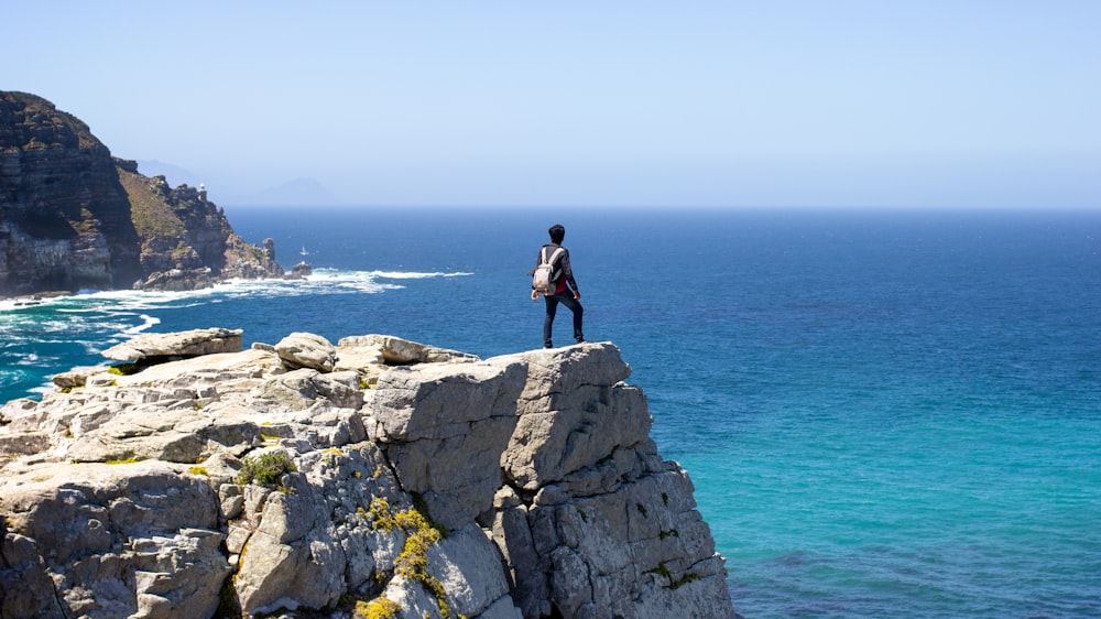 uomo in giacca nera in piedi sulla formazione rocciosa grigia vicino allo specchio d'acqua durante il giorno