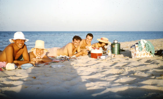 A joyful group of travelers laughing together on a sunlit beach with turquoise ocean waves behind them.