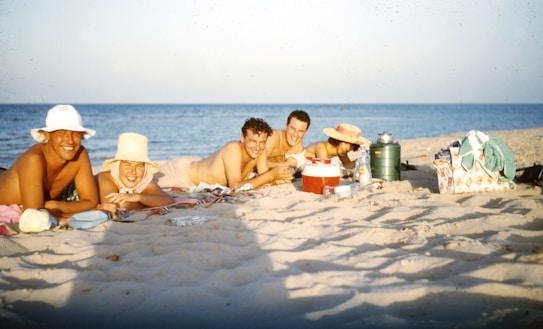 A group of people relaxing and smiling on a sandy beach. They are lying on towels and surrounded by beach gear, such as coolers, a thermos, and a beach bag. The ocean appears in the background under a clear blue sky.