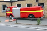 A large red fire truck with yellow stripe details is parked beside a brick building. On the pavement in front of the vehicle lies a person in an orange and beige uniform, appearing to be a firefighter. There is a fence in the foreground separating the sidewalk from the vehicle area.