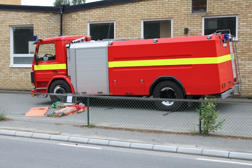 A large red fire truck with yellow stripe details is parked beside a brick building. On the pavement in front of the vehicle lies a person in an orange and beige uniform, appearing to be a firefighter. There is a fence in the foreground separating the sidewalk from the vehicle area.