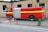 A large red fire truck with yellow stripe details is parked beside a brick building. On the pavement in front of the vehicle lies a person in an orange and beige uniform, appearing to be a firefighter. There is a fence in the foreground separating the sidewalk from the vehicle area.