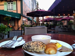 A quaint outdoor café setting features a table with a plate of pastries, including a sesame seed bun and a croissant, accompanied by a small bowl of butter and sliced meat. The café is surrounded by charming buildings with green shutters, and purple umbrellas provide shade over other tables. The atmosphere is peaceful with lush green plants adding to the cozy ambiance.