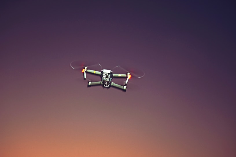 A drone with four rotors is flying against a gradient sky transitioning from deep blue to warm orange. The drone's rotors are blurred due to motion, and its body appears to be metallic with some colored elements.