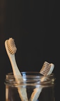 Two bamboo toothbrushes are placed inside a glass jar against a dark background. The toothbrushes feature natural wood handles and bristles, emphasizing an eco-friendly design.