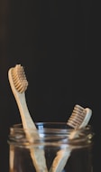 Two bamboo toothbrushes are placed inside a glass jar against a dark background. The toothbrushes feature natural wood handles and bristles, emphasizing an eco-friendly design.