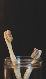 Two bamboo toothbrushes are placed inside a glass jar against a dark background. The toothbrushes feature natural wood handles and bristles, emphasizing an eco-friendly design.