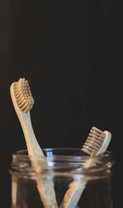 Two bamboo toothbrushes are placed inside a glass jar against a dark background. The toothbrushes feature natural wood handles and bristles, emphasizing an eco-friendly design.
