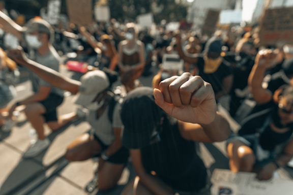 A group of people kneeling with raised fists during a protest. Most are wearing masks and appear engaged in a collaborative action, with signs visible in the background and bright sunlight casting shadows on the ground.