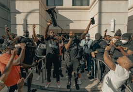 A group of people gather outside a building, many holding phones or cameras. One person stands prominently in the center holding a megaphone aloft, with other individuals around them wearing masks. The atmosphere is charged and communal.