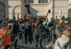A group of people gather outside a building, many holding phones or cameras. One person stands prominently in the center holding a megaphone aloft, with other individuals around them wearing masks. The atmosphere is charged and communal.