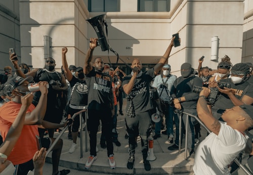 A group of people gather outside a building, many holding phones or cameras. One person stands prominently in the center holding a megaphone aloft, with other individuals around them wearing masks. The atmosphere is charged and communal.