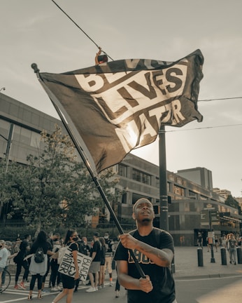 A person is holding a large flag that reads 'Black Lives Matter' amidst a group of people gathered in an urban area. Some individuals are holding signs and many of them appear to be participating in a protest or demonstration. Modern buildings and trees are visible in the background under a clear sky.