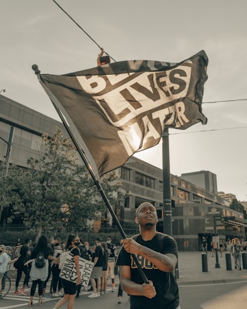 A person is holding a large flag that reads 'Black Lives Matter' amidst a group of people gathered in an urban area. Some individuals are holding signs and many of them appear to be participating in a protest or demonstration. Modern buildings and trees are visible in the background under a clear sky.