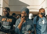A group of individuals stands in front of a building, with a central figure holding a megaphone. The person in the middle wears glasses and a headwrap, and appears to be speaking. The others are dressed in a variety of clothing, some with printed messages on their shirts.