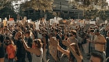 A large crowd of people engaged in a protest, holding up various signs and banners with messages. The participants appear diverse in age and ethnicity, and many are wearing masks. The scene is set outdoors against the backdrop of urban buildings, and the mood appears to be one of solidarity and activism.