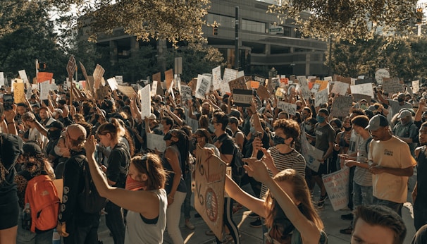 A large crowd of people engaged in a protest, holding up various signs and banners with messages. The participants appear diverse in age and ethnicity, and many are wearing masks. The scene is set outdoors against the backdrop of urban buildings, and the mood appears to be one of solidarity and activism.