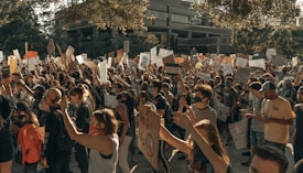 A large crowd of people engaged in a protest, holding up various signs and banners with messages. The participants appear diverse in age and ethnicity, and many are wearing masks. The scene is set outdoors against the backdrop of urban buildings, and the mood appears to be one of solidarity and activism.