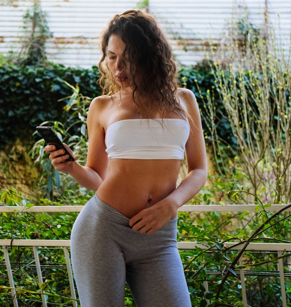 woman in white tank top and gray pants holding black smartphone