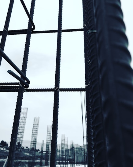 Metal rebar structures stand vertically, forming a framework against a pale sky. In the background, additional rebar formations are visible, likely part of a construction site. The image conveys a sense of industrial progress and development.